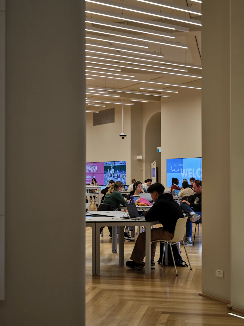Students studying with laptops in bright modern library, warm natural light streaming through large windows